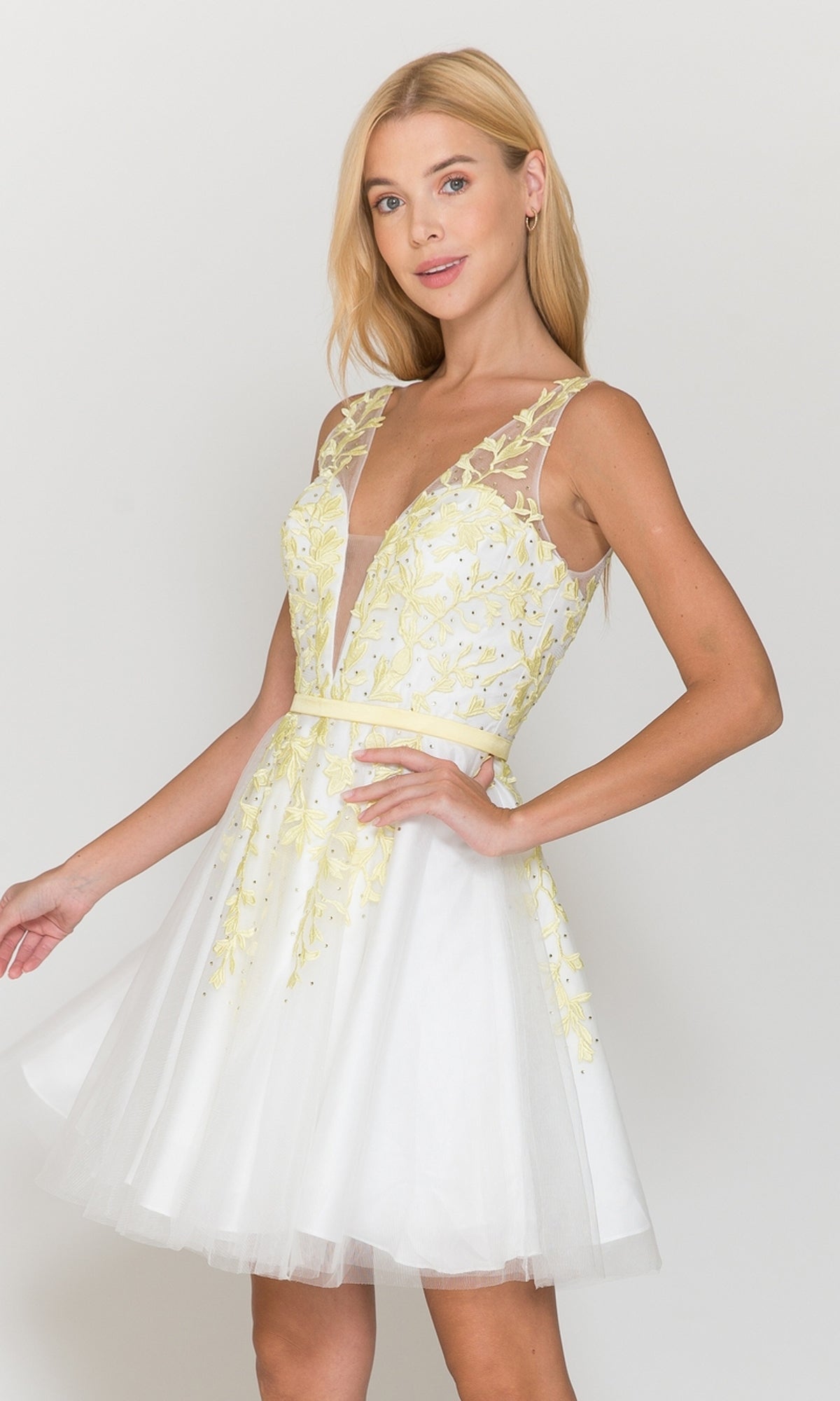 A woman wears the Tulle and Lace Short A-Line Homecoming Dress 8696 in White/Yellow, featuring a sleeveless design, sheer deep V neckline, yellow floral embroidery, and stands against a plain light background.